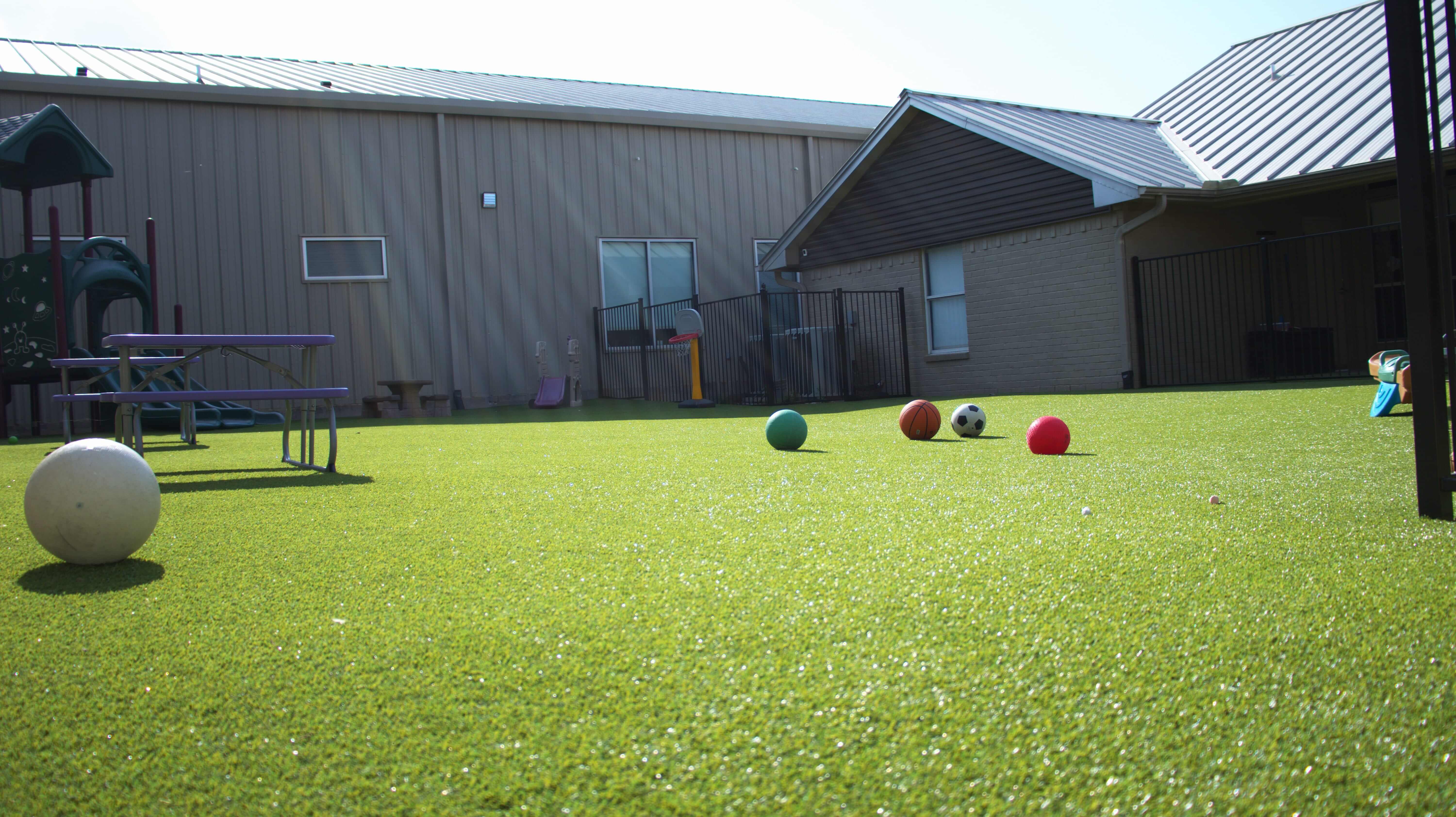 Play area with artificial grass, sports equipment, and a slide near a building with a metal roof